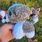 Hand holding a small plush toy resembling a creature with a textured body and white feet against a grassy background.